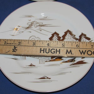 May include: Set of six white ceramic plates with a gold rim and a Japanese-inspired design featuring a mountain, trees, and a house. The plates are 6 inches in diameter and are made in the USA.