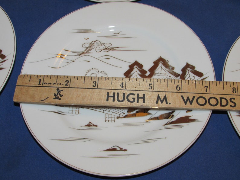 May include: Set of six white ceramic plates with a gold rim and a Japanese-inspired design featuring a mountain, trees, and a house. The plates are 6 inches in diameter and are made in the USA.