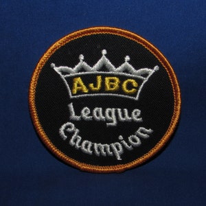 May include: A black and gold embroidered patch with a crown and the text "AJBC League Champion".