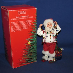 May include: A Santa Claus figurine wearing a white jacket with red patterned suspenders and red trousers, tangled in a string of multicolored Christmas lights. Beside the figurine is a tall red box labeled Possible Dreams Santa's Handwork.