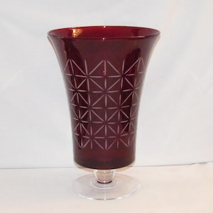 May include: A tall, burgundy-colored glass vase with a clear, circular base. The vase has a flared top and is decorated with a geometric starburst pattern. The vase is approximately 10 inches tall.