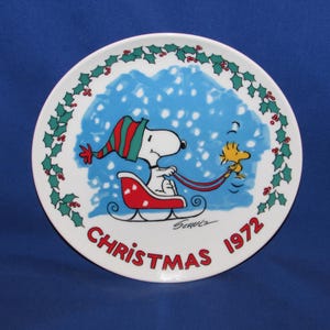 May include: A round, white Christmas plate featuring Snoopy in a red sled, wearing a green and red hat, with Woodstock. The plate has a blue snowy background and a holly berry border. The text "CHRISTMAS 1972" is in red.