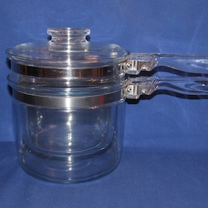 May include: A clear glass double boiler with a lid and two clear handles. The pot has a silver band around the middle and a clear glass lid with a small knob. The handles are clear and attached to the side of the pot.