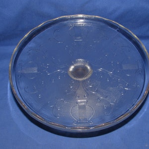 May include: A clear glass cake stand with a decorative etched design of a stylized urn with floral accents. The stand has a scalloped edge.