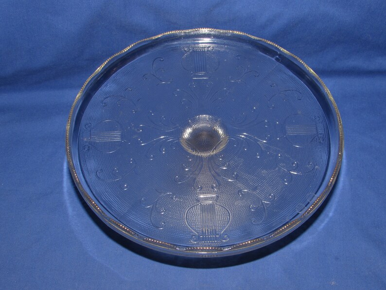 May include: A clear glass cake stand with a decorative etched design of a stylized urn with floral accents. The stand has a scalloped edge.