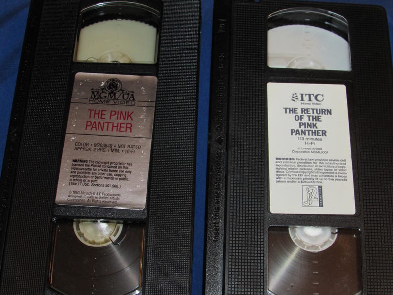 May include: Two black VHS tapes featuring the movies "The Pink Panther" and "The Return of the Pink Panther." The tapes have white labels with movie titles and logos. The tapes are in black plastic cases.