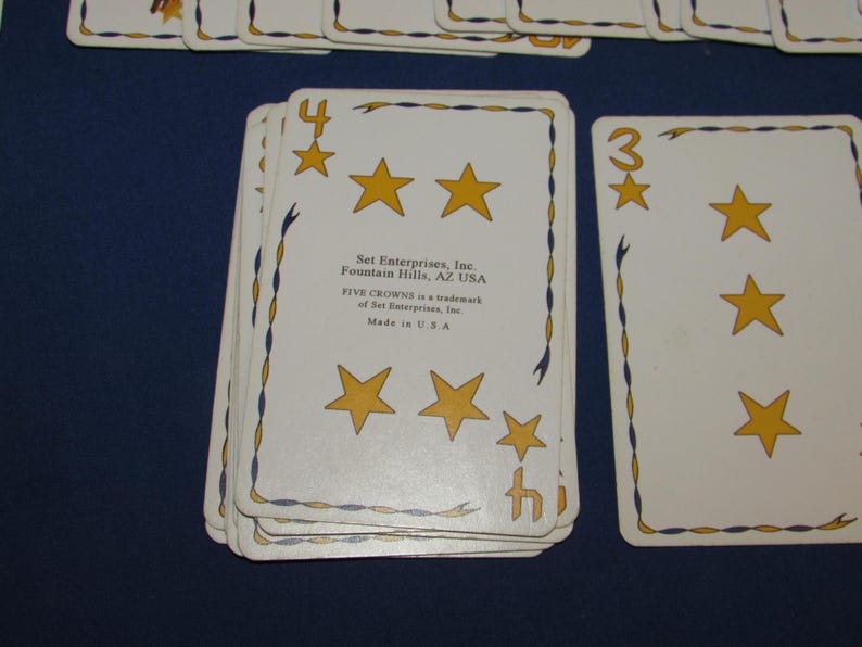 May include: A deck of Five Crowns playing cards. The cards are white with gold stars and blue and gold decorative borders. The number 4 and 3 are visible on the cards.
