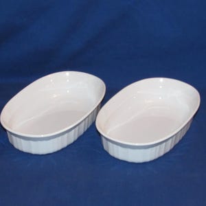 May include: Two identical oval-shaped white ceramic baking dishes with fluted sides. Each dish features a smooth interior basin and a clean, glossy finish, designed for oven use with a consistent depth and a simple, functional rim structure.