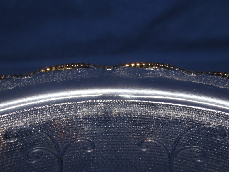 May include: A clear glass serving dish with a scalloped edge and a gold rim. The dish has a textured surface with a repeating pattern of small dots and swirls.