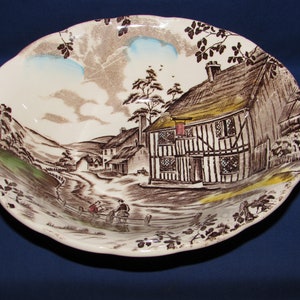 May include: A brown and white oval ceramic bowl with a scene of a village with a brown and white building in the center. The bowl has a scalloped edge and a brown and white floral design.