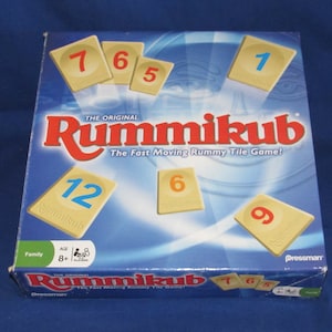 May include: The Original Rummikub game box with tiles. The box is blue with red and white text. Several tiles with numbers 1, 5, 6, 7, 9, and 12 are visible. The game is for ages 8+.