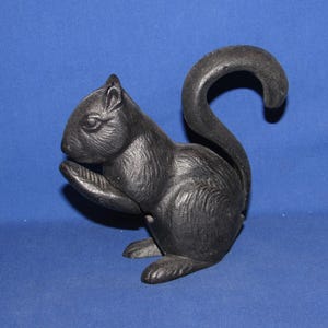May include: A black cast iron nutcracker shaped like a squirrel. The squirrel is depicted in a seated position with its tail curved upward and one paw raised toward its face. The surface features textured details representing fur and facial features.