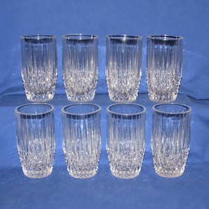 May include: Eight clear, cut-glass tumblers arranged in two rows on a blue surface. The glasses have a vertical ribbed design and a diamond-patterned base. The rims are smooth and the glasses are empty.