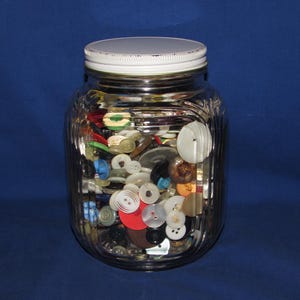 May include: A clear glass jar filled with a variety of colorful buttons. The jar has a white lid and a ribbed design. The buttons are in various sizes, shapes, and colors, including red, white, black, and blue.