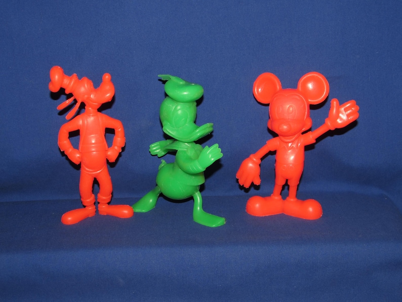 May include: Three plastic cartoon character figurines. A red Goofy, a green Donald Duck, and a red Mickey Mouse.