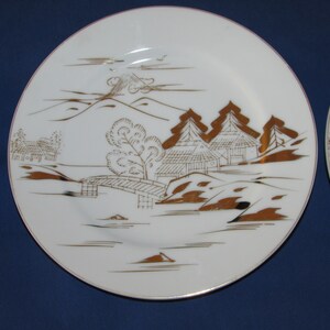 May include: A white ceramic plate with a gold rim and a Japanese-inspired design. The design features a mountain, trees, houses, and a bridge, all in gold.