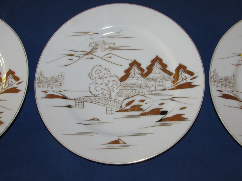 May include: A white ceramic plate with a gold rim and a Japanese-inspired design. The design features a mountain, trees, houses, and a bridge, all in gold.