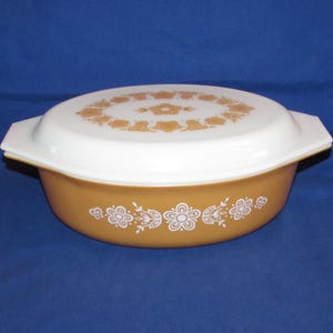 May include: An oval-shaped, amber-colored casserole dish with a white lid. The dish features a floral pattern in white and amber. The lid has a central floral design and a border of flowers.