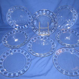 May include: Eight clear glass dinner plates with a scalloped edge. The plates are arranged in a circle on a blue background.
