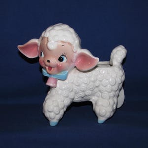 May include: A white ceramic lamb planter featuring a textured curly wool design, large expressive eyes, pink-tinted ears and cheeks, and a light blue bow tied around its neck. The lamb has a small opening on its back for holding small plants.