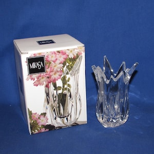 May include: A clear, decorative glass vase with a unique, pointed rim design. The vase is next to its original box, which features the Mikasa logo and a floral arrangement. The vase is approximately 7 inches tall.
