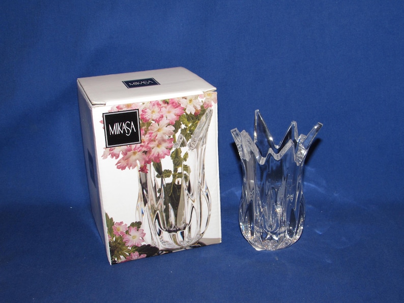 May include: A clear, decorative glass vase with a unique, pointed rim design. The vase is next to its original box, which features the Mikasa logo and a floral arrangement. The vase is approximately 7 inches tall.
