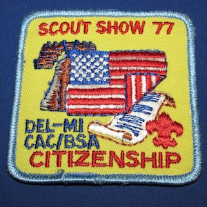 May include: A yellow embroidered patch with blue edging. The patch features a red, white, and blue American flag, a scroll, and a fleur-de-lis. The text on the patch reads "Scout Show 77 Del-Mi CAC/BSA Citizenship".