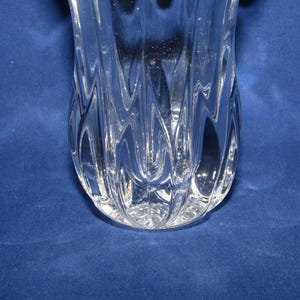 May include: A clear glass vase with a textured, wave-like design. The vase has a wide, rounded base and tapers upwards. The glass is transparent, allowing light to pass through, and is set against a blue background.