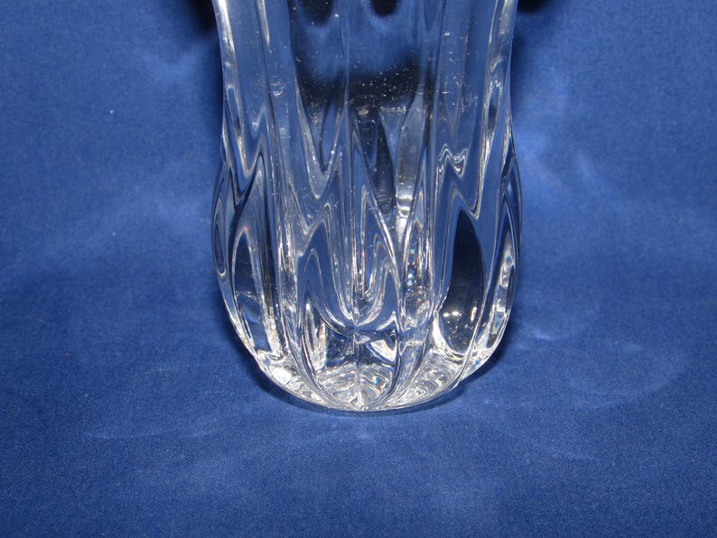 May include: A clear glass vase with a textured, wave-like design. The vase has a wide, rounded base and tapers upwards. The glass is transparent, allowing light to pass through, and is set against a blue background.