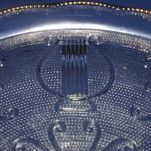 May include: A clear glass plate with a gold rim and a raised design of a lyre in the center. The plate has a textured surface.