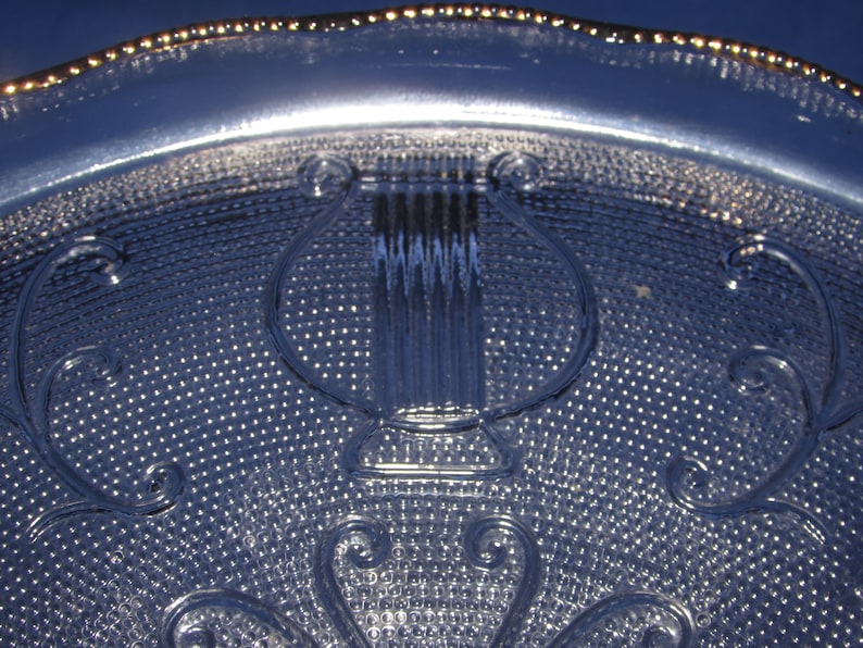 May include: A clear glass plate with a gold rim and a raised design of a lyre in the center. The plate has a textured surface.