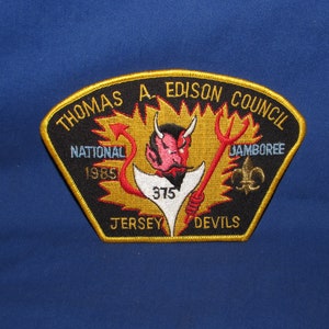 May include: A black and gold embroidered patch with the text "Thomas A. Edison Council National 1985 Jamboree Jersey Devils" and a red devil holding a pitchfork.