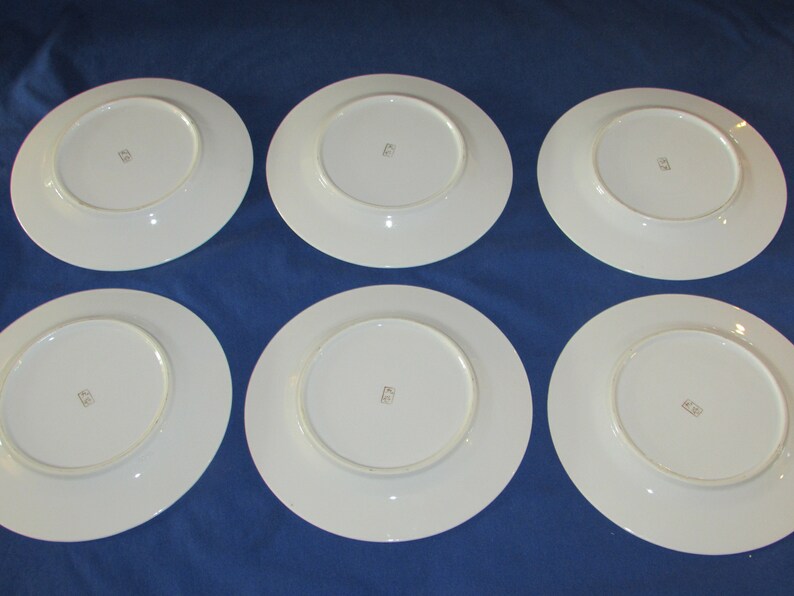 May include: Six white ceramic dinner plates with a gold rim and a small gold mark on the bottom.