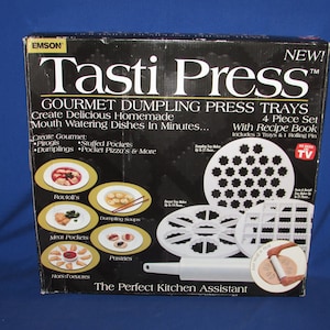 May include: A black and white box with the words "Tasti Press" in white. The box features a set of four white plastic trays for making dumplings, pierogies, and other filled pastries. The box also includes a rolling pin and a recipe book. The text on the box reads "Gourmet Dumpling Press Trays, Create Delicious Homemade Mouth Watering Dishes In Minutes... 4 Piece Set With Recipe Book Includes 3 Trays & 1 Rolling Pin".