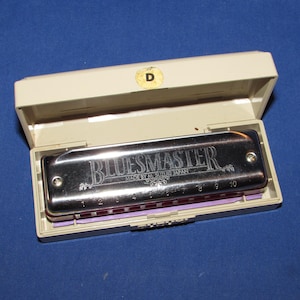 May include: A silver Bluesmaster harmonica in an open beige case. The harmonica has the word "BLUESMASTER" engraved on it, along with the text "MADE BY A. SUZUKI JAPAN". The case is rectangular with a purple interior.