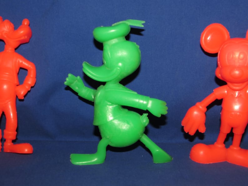 May include: Three vintage plastic cartoon character figurines. A red Goofy, a green Donald Duck, and a red Mickey Mouse.
