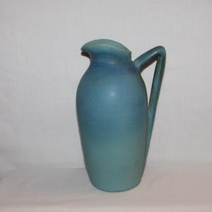 May include: A ceramic pitcher featuring a matte finish with a gradient color scheme that transitions from a deep, dark blue at the top to a lighter, pale blue at the base. The vessel has a curved handle and a simple, rounded pouring spout.