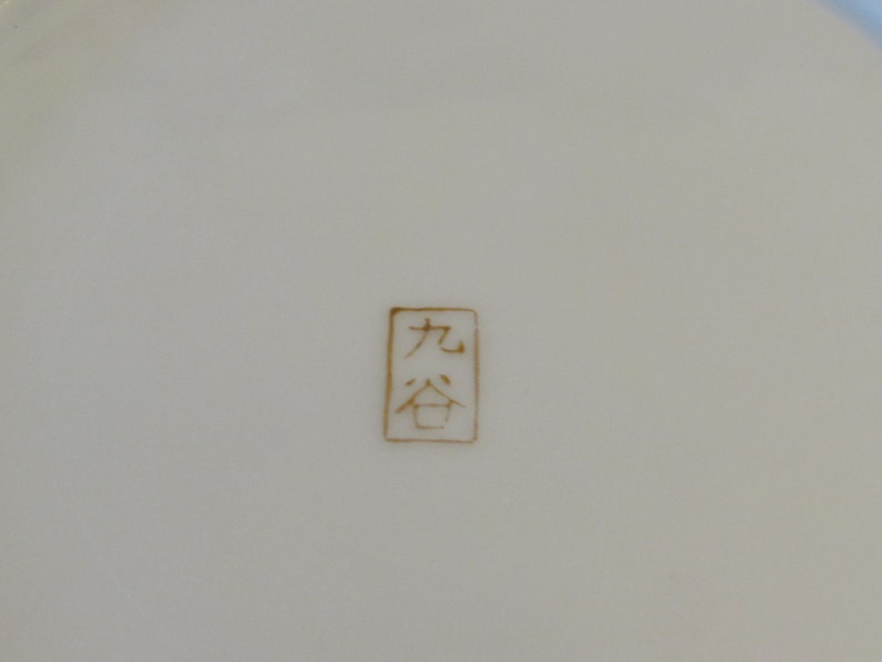 May include: A close-up of a white ceramic plate with a gold stamp that reads "九谷".