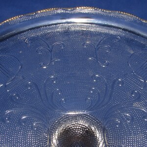 May include: A clear glass cake stand with a gold rim and a decorative pattern of stylized harps and swirls. The stand has a textured surface.