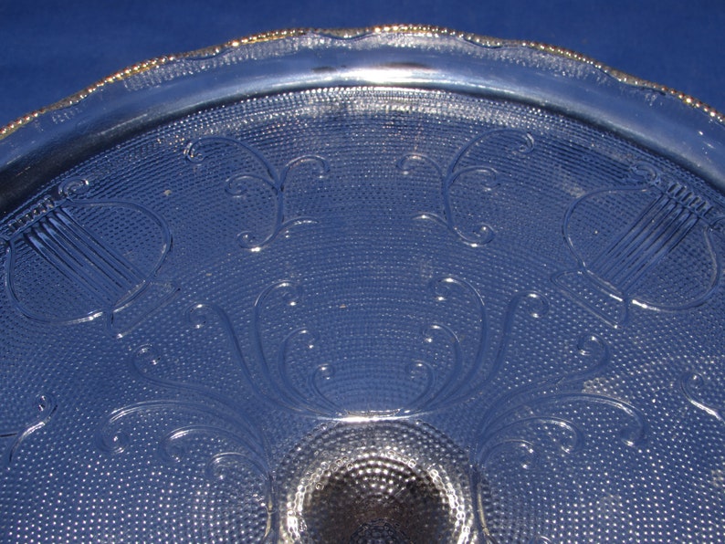 May include: A clear glass cake stand with a gold rim and a decorative pattern of stylized harps and swirls. The stand has a textured surface.