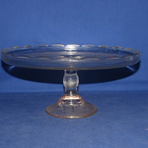 May include: A clear glass cake stand with a scalloped edge and a pedestal base. The stand has a delicate etched design.