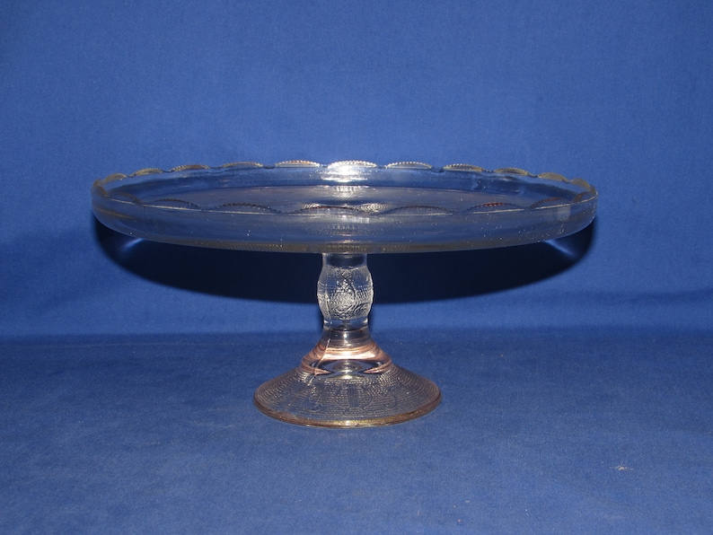 May include: A clear glass cake stand with a scalloped edge and a pedestal base. The stand has a delicate etched design.