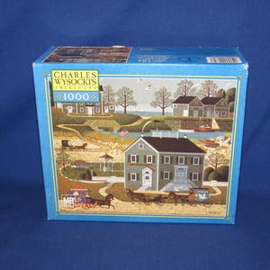 May include: A 1000 piece jigsaw puzzle box featuring a painting by Charles Wysocki. The box shows a quaint town scene with houses, trees, and a horse-drawn carriage. The title of the painting is "American" and the puzzle is made by MB.