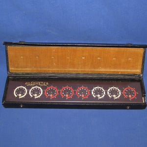 May include: A vintage Addometer mechanical adding machine housed in a black case with a tan interior. The device features a dark brown faceplate with eight circular dials, each displaying white and red numerical markings for manual calculation.