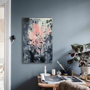 May include: A desk with a wooden top and legs, a gray throw blanket, a magazine, a white candle, and a plant in a white pot. A painting with pink, white, and black abstract shapes hangs on the wall behind the desk.