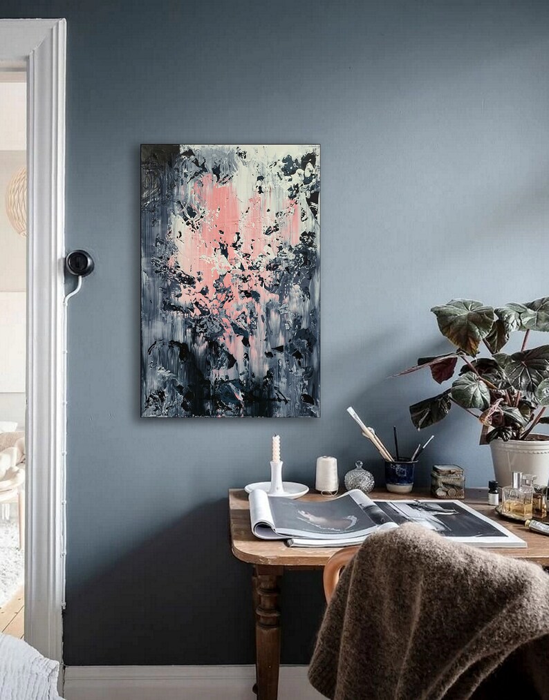 May include: A desk with a wooden top and legs, a gray throw blanket, a magazine, a white candle, and a plant in a white pot. A painting with pink, white, and black abstract shapes hangs on the wall behind the desk.