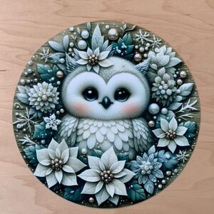 May include: A white owl with large, dark eyes is surrounded by white poinsettia flowers and green leaves. The owl is sitting in the center of a round, wooden frame. The background is a light brown wood.