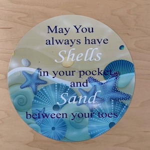 May include: A round, blue and white sign with a seashell and starfish design. The sign reads "May You always have Shells in your pocket and Sand between your toes."