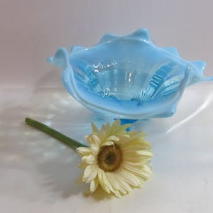 May include: A light blue glass bowl with a ruffled edge, resembling a seashell. A yellow and white daisy with a green stem rests in front of the bowl. The bowl has a textured interior.