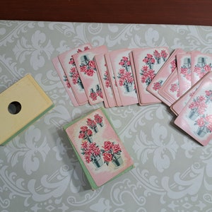May include: A vintage deck of playing cards with a floral design. The cards have a pink background and feature a silver bucket filled with red flowers. A light yellow card box with a hole is also present.